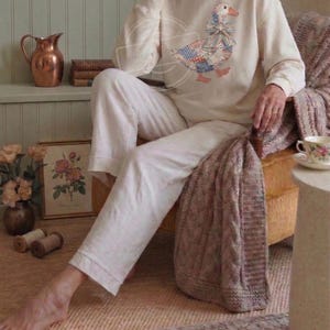 May include: Cream-colored sweatshirt with a patchwork goose design, paired with white pants. The goose features a blue, red, and white pattern. The image has a cozy, vintage aesthetic, suggesting loungewear or casual apparel.