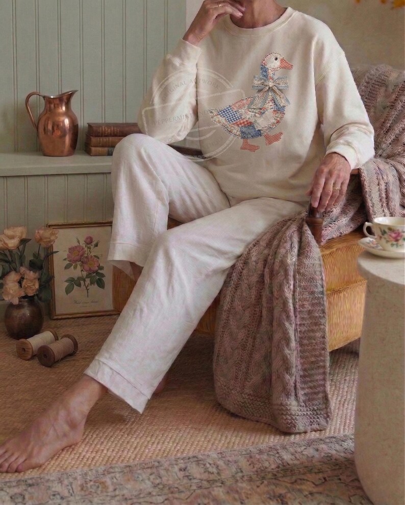 May include: Cream-colored sweatshirt with a patchwork goose design, paired with white pants. The goose features a blue, red, and white pattern. The image has a cozy, vintage aesthetic, suggesting loungewear or casual apparel.