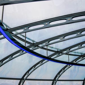 Puede incluir: Detalle arquitectónico moderno con una estructura metálica negra curvada y un acento azul. El diseño incluye vigas paralelas con cortes ovalados, que soportan un techo transparente. Estética contemporánea.