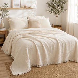May include: A cream-coloured textured bedspread with a fringed edge draped over a bed. The bedding set includes two large rectangular pillows and one smaller square pillow in a matching cream and light beige tone, creating a layered look.