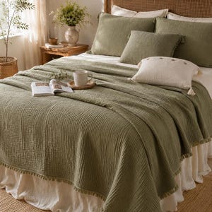 May include: A sage green crinkled cotton bedspread with fringe trim, featuring two matching sage green pillow shams, one square sage green throw pillow, and one rectangular cream-colored throw pillow with button accents on a bed.
