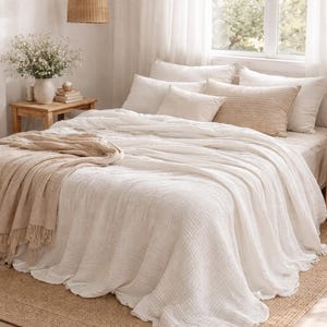 Può includere: Un copriletto bianco stropicciato con un delicato bordo in pizzo, sovrapposto a una coperta beige testurizzata. Il letto è decorato con diversi cuscini bianchi e beige, incluso uno con una trama intrecciata, su una struttura letto in legno.