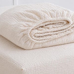 May include: A folded off-white fitted sheet with a crinkled, textured surface. The fabric features a gathered elastic edge along the perimeter, creating a soft, layered appearance. The material has a consistent, light cream color throughout.