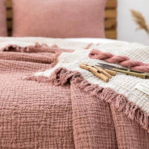 Könnte beinhalten: Eine wendbare Tagesdecke mit texturiertem Waffelmuster in Altrosa und Creme. Die Decke hat ausgefranste Ränder und ein kleines weißes Stoffetikett mit Text. Ein Strang rosafarbenes Garn, hölzerne Wäscheklammern und eine Schere liegen darauf.