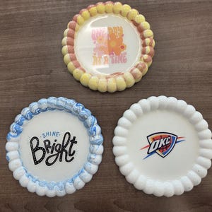 Può includere: Tre sottobicchieri decorativi in ceramica con bordi smerlati. Uno ha il testo "ONE DAY AT A TIME" con accenti floreali. Un altro dice "SHINE. Bright". Il terzo presenta il logo OKC Thunder.