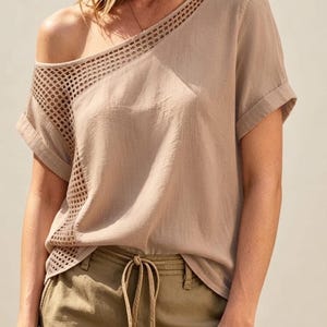 Puede incluir: Una blusa beige de manga corta con un escote ancho y asimétrico y un panel diagonal de tela de malla abierta que se extiende desde el hombro hasta el costado. La prenda tiene mangas con dobladillo y se combina con pantalones verde oliva con cordón.