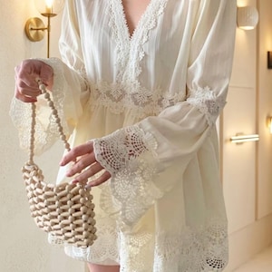 May include: Cream-colored dress with lace trim and long sleeves. The dress features a V-neckline and a tiered skirt. A small, beaded wooden handbag is held in the hand.