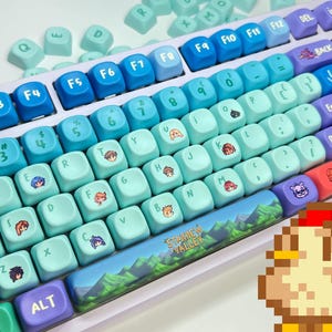 以下が含まれることがあります： グラデーションの青とティール色のキーキャップが付いたカスタムキーボード。キーにはキャラクターアートと「Stardew Valley」の文字が描かれています。キーボードは白いフレームとピクセル化された鶏のグラフィックを備えています。