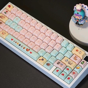 以下が含まれることがあります： 白枠のパステルカラーのメカニカルキーボード。キーキャップはピンク、ブルー、イエローで、様々なピクセルアートのデザインとシンボルが施されています。小さなフィギュアが横にあります。キーボードは標準的なQWERTYレイアウトです。