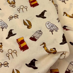 May include: A cream-colored fabric featuring a repeating pattern of wizarding-themed illustrations including white owls, brown sorting hats, red and yellow striped scarves, gold snitches, birdcages, round glasses, and quidditch hoops.