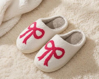 Può includere: Un paio di pantofole bianche e soffici con un design a fiocco rosa ricamato sulla parte superiore. L'interno è grigio chiaro e le pantofole sono appoggiate su una superficie strutturata color crema.