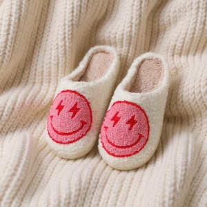 Könnte beinhalten: Ein Paar weiße Plüschhausschuhe mit einem rosa Smiley-Gesicht-Design und roten Blitz-Akzenten. Die Hausschuhe haben ein weiches, hellrosa Innenfutter und liegen auf einer cremefarbenen Strickoberfläche.