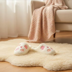 May include: A pair of white, embroidered slippers with pink seashell and flower designs. The slippers are on a cream-colored, textured rug, with a beige chair and a pink blanket in the background. The slippers appear soft and comfortable.