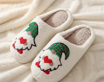 Puede incluir: Un par de zapatillas de felpa blancas con un diseño de gnomo. Los gnomos tienen sombreros verdes y rojos, narices rojas en forma de corazón y contornos negros. Las zapatillas tienen un interior suave y difuso y están sobre una superficie blanca texturizada.