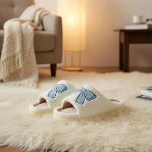 May include: A pair of cream-coloured, fluffy slippers featuring light blue bow embroidery. The slippers have a soft, beige interior and are placed on a cream-coloured, shaggy rug. The background includes a beige sofa, a floor lamp, and a small wooden table.