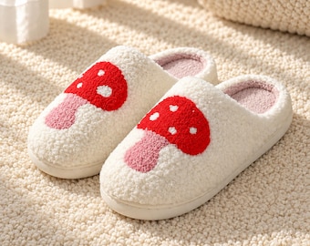 Può includere: Un paio di pantofole slip-on color crema, soffici e pelose, con un grafico a forma di fungo rosso e rosa sulla parte superiore. Le pantofole presentano un esterno testurizzato tipo sherpa, una fodera interna rosa chiaro e una suola solida.