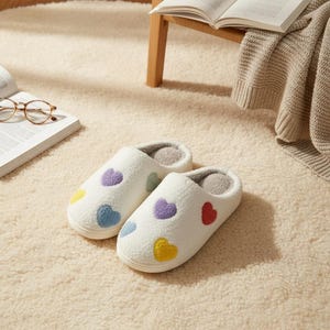 Könnte beinhalten: Ein Paar weiße, flauschige Hausschuhe mit bunten, gestickten Herzen. Die Hausschuhe haben ein weiches, graues Innenfutter. Die Herzen sind gelb, blau, lila und rot. Für den Innenbereich.