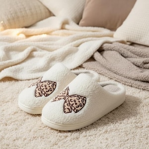 Op de afbeelding: Een paar crèmewitte, zachte pantoffels met een crèmekleurige zool. Elke pantoffel heeft een geborduurde strik met een bruin en beige luipaardprint. De pantoffels liggen op een crèmekleurig tapijt, met kussens en dekens op de achtergrond.