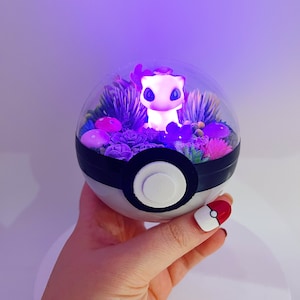 May include: A clear glass dome with a purple and white Pokemon character inside. The dome is filled with dried flowers and greenery and sits on a black and white Pokeball base.