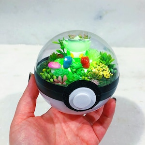 May include: A clear glass dome with a black and white Pokeball base. Inside the dome is a miniature green and white Pokemon character surrounded by green and yellow plants.