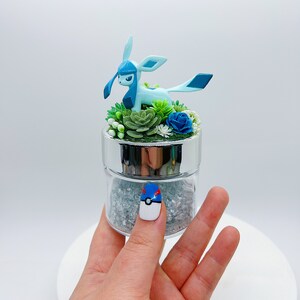 May include: A glass jar with a silver lid filled with clear glitter and a small blue and white Pokemon figurine. The figurine is surrounded by green and blue artificial flowers.
