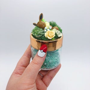 Op de afbeelding: Een klein glazen potje met een koperen deksel gevuld met groene glitter en een klein, slapend bruin en beige Pokémon-figuurtje omringd door groene en witte bloemen.