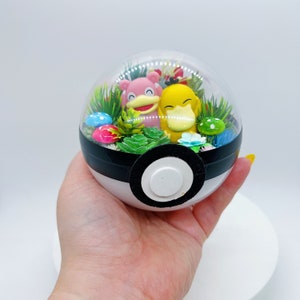 Puede incluir: Un terrario de Poke Ball transparente con una figura de Pokemon rosa y amarilla en su interior. El terrario está lleno de hierba verde, flores de colores y pequeños champiñones.