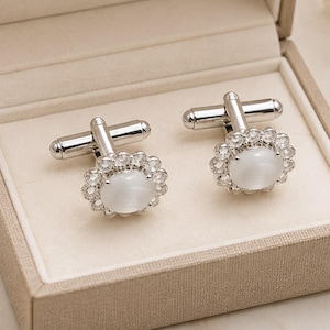 May include: A pair of silver-toned cufflinks featuring a central oval white cabochon stone surrounded by a halo of small clear crystals. The cufflinks are presented inside a beige fabric-covered box with a hinged lid and a cream-colored interior.