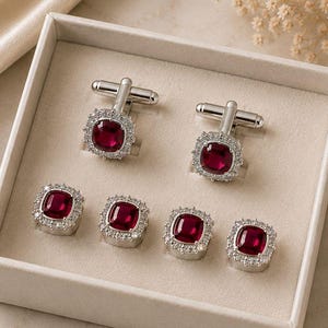 May include: A set of silver cufflinks and shirt studs displayed in a white box. Each piece features a square, deep red gemstone surrounded by a halo of smaller, clear stones. The set includes two pairs of cufflinks and three shirt studs.