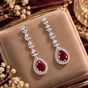 Puede incluir: Un par de pendientes largos con una fila vertical de tres piedras transparentes en forma de pera que terminan en una piedra roja oscura más grande en forma de pera, rodeada por un halo de pequeñas piedras transparentes en metal plateado.