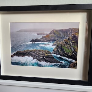 May include: Framed print of a coastal landscape, showing cliffs, ocean waves, and a misty sky. The print is mounted with a white mat and a black wooden frame. The sea is a vibrant turquoise colour.