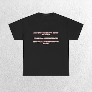 May include: Black t-shirt with the text "ZERO EPISODES OF LOVE ISLAND WATCHED, ZERO DUBAI CHOCOLATE EATEN, ZERO ONLYFANS SUBSCRIPTIONS BOUGHT" in pink lettering. The shirt is a basic crew neck style.