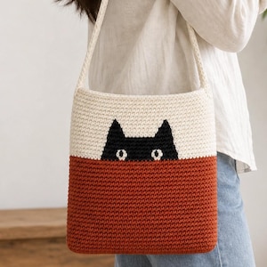 May include: A rectangular crochet shoulder bag featuring a two-tone design with a cream-colored upper section and a rust-orange lower section. A black cat face with white eyes is stitched onto the cream portion of the bag, which has a long strap.