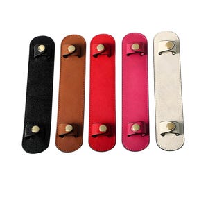 May include: Five leather shoulder strap pads in black, brown, red, pink, and white. Each pad has a gold-coloured snap closure. The pads are rectangular and designed to protect the shoulder from the weight of a bag or purse.