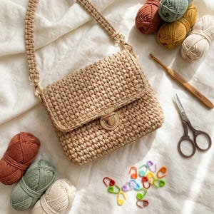 Könnte beinhalten: Beige gehäkelte Handtasche mit goldenem Verschluss und Riemen, umgeben von Garn in verschiedenen Farben, einem Häkelhaken, einer Schere und Maschenmarkierern. Die Tasche ist rechteckig mit einem Klappenverschluss.