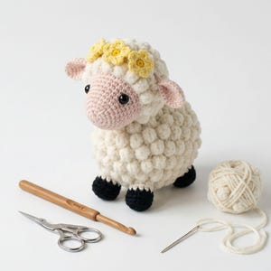 Puede incluir: Una oveja de ganchillo hecha a mano con cuerpo color crema, cara y orejas rosas y pezuñas negras. La oveja lleva una corona de flores amarillas. Un ganchillo, tijeras, hilo y una aguja están junto a la oveja.