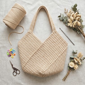 May include: A beige crochet tote bag with a geometric design, alongside a ball of yarn, scissors, stitch markers, and a crochet hook. Dried flowers and greenery add a decorative touch to the scene. The bag has two handles.