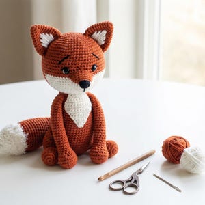 Puede incluir: Muñeco de zorro tejido a ganchillo en tonos naranja, blanco y negro. El zorro tiene el pecho y la punta de la cola blancos, con ojos y nariz negros. Suministros de ganchillo, incluyendo hilo, un ganchillo, tijeras y una aguja, están cerca.