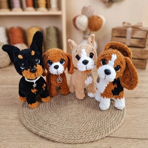 以下が含まれることがあります： 黒、茶色、白の手作りの犬の編みぐるみ4体。それぞれユニークなデザインで、足跡のチャームが付いているものもあります。丸い織りのマットの上に配置されています。 Etsyのロゴが左下に表示されています。
