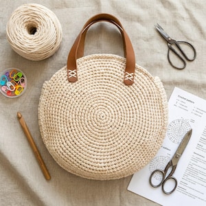 May include: A cream-colored, circular crochet bag with brown leather handles and white stitching. The bag is surrounded by crochet supplies, including a ball of yarn, a crochet hook, stitch markers, and scissors. A crochet pattern is also visible.