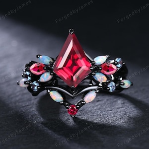 May include: A black metal ring featuring a central kite-shaped red gemstone, accented with smaller marquise-cut opals and round red stones. The band has a V-shaped design, and the text ForgedbyJames is visible in the background.