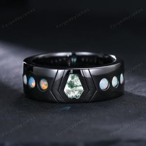 May include: A black ring with a central geometric design featuring a green moss agate stone. The ring is accented with opal stones and has the text "ForgedbyJames" repeated around the band.