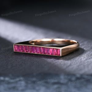 May include: A rose gold ring featuring a horizontal bar set with a row of vibrant, rectangular-cut pink rubies. The band has a smooth, polished finish, and the stones are arranged in a channel setting across the top of the ring's flat surface.