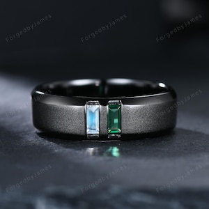 Puede incluir: Un anillo negro con una banda texturizada, con dos piedras rectangulares: una azul claro y otra verde esmeralda. El anillo se encuentra sobre un fondo oscuro, resaltando el contraste de colores y texturas.