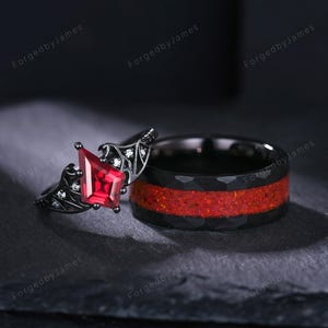 Puede incluir: Dos anillos: un anillo de compromiso negro con una piedra preciosa roja y pequeñas piedras blancas, y una alianza de boda negra con un centro rojo. Los anillos están sobre un fondo oscuro.