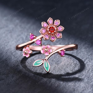 May include: A rose gold bypass ring featuring a floral design with a central red stone surrounded by pink stones, smaller pink stone accents, and two light green enamel leaves. The ring has an open, adjustable band structure with delicate metal stems.
