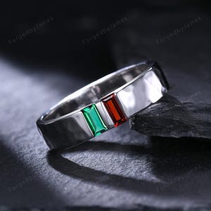 May include: A silver ring with a hammered texture. The ring features three rectangular gemstones in green, white, and red, arranged in a row. The ring is displayed on a dark, textured surface.