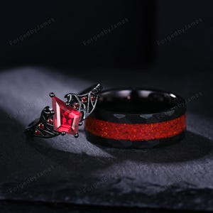 May include: Two black rings with red detailing. One ring has a large, red, kite-shaped gemstone and smaller red stones. The other ring features a red band set in black metal. The rings are displayed on a dark surface.