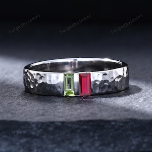 May include: A silver-toned metal ring featuring a hammered, textured surface. The band is set with two rectangular, baguette-cut gemstones positioned side-by-side: one light green peridot and one deep red ruby, both oriented vertically.