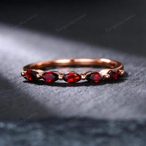 May include: A rose gold ring featuring a series of marquise-cut, deep red gemstones. The gemstones are set closely together, creating a continuous band of color. The ring is displayed on a dark, textured surface.